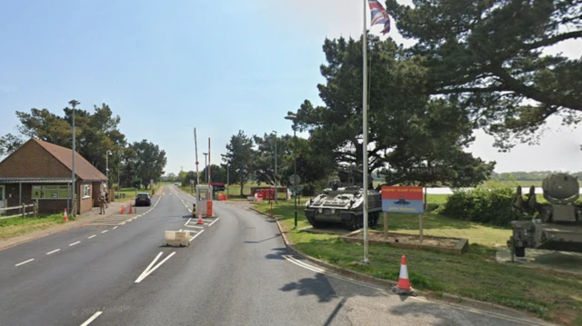 A screenshot from Google street view showing the entrance to Thorney Island military base. There is a one-storey shed on the left with a man in uniform holding a gun. On the right there are two green tanks, a sign and a flagpole.