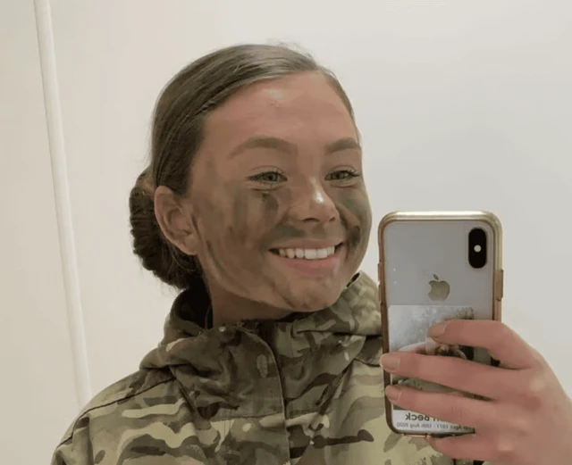 A mirror selfie of Jaysley Beck wearing her camouflage formal military uniform and paint on her face. She has her brown hair tied into a tight bun and is smiling at the camera.