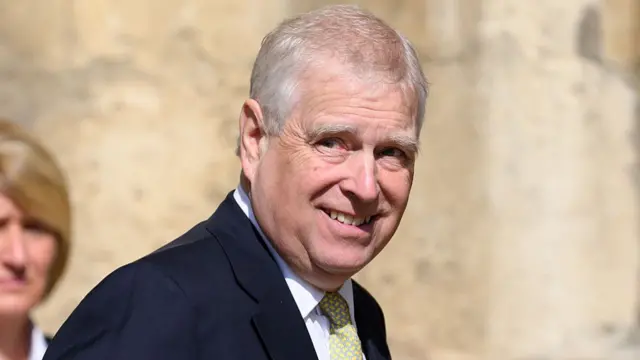 A man with grey hair looking at the camera and smiling, head slightly turned. He is wearing a blue suit with a green and yellow tie.