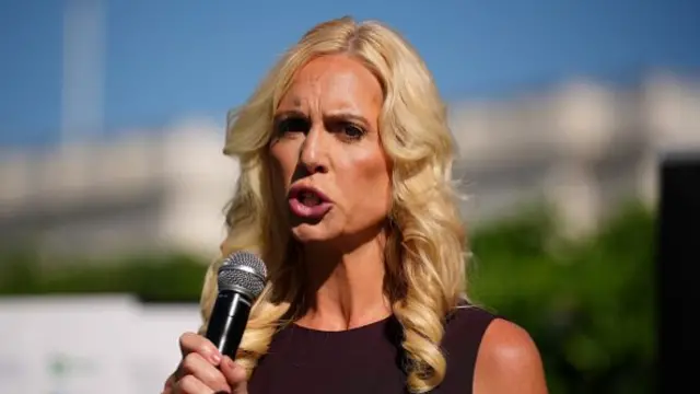 A woman with blonde curled hair speaks holding a microphone