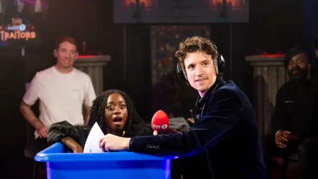 Greg James, wearing headphones, looks around as a woman pops out of a blue tub.