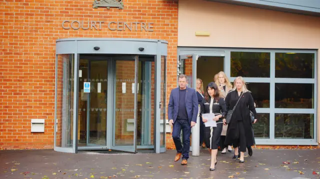 A family of three women and a man stand walking out of a court speaking to members of the press. A woman is front and centre and holding a piece of paper. They are all smartly dressed.