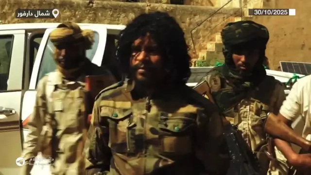 Footage showing Abu Lulu being detained by RSF fighters. Two armed men stand behind Abu Lulu as he is led away from a truck.