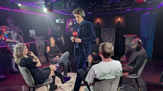 Group of people sat in circle on grey chairs, with man in blue suit holding mic standing in middle