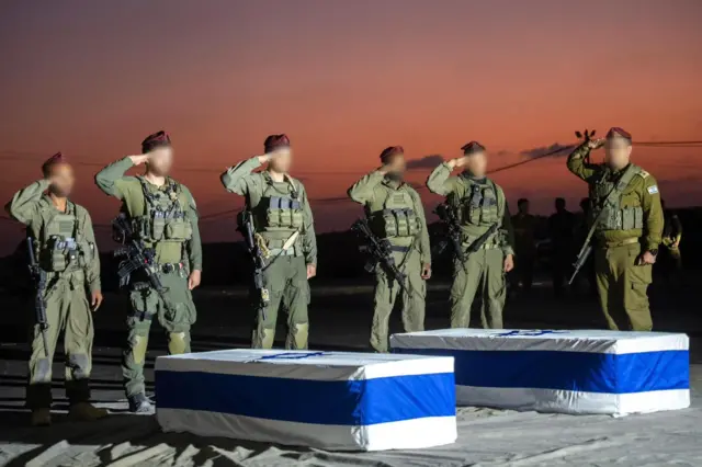 Five IDF soldiers stand to attention and salute coffins draped in Israeli flags.