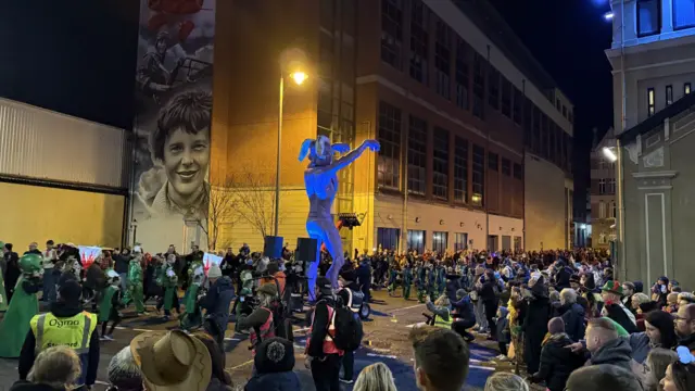 People gather in the street for the parade as a large figure passes