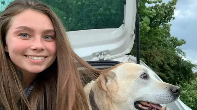 A selfie of Jaysley Beck sitting in the open boot of her car next to a white Labrador.