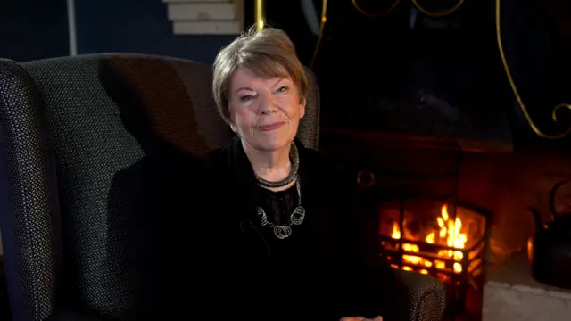 A woman is smiling at the camera, she is sat in a high armchair in front of an open fire