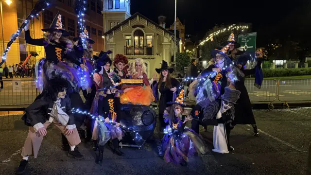 Fans have dressed up as hocus pocus characters as they surround a black cauldron, with orange lights imitating a boiling cauldron. The girls are surrounded by people wearing black costumes with witches hats, and broomsticks with led lights attached