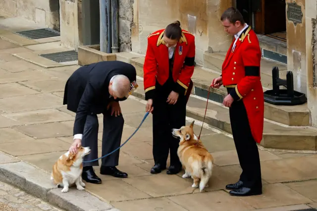 Andrew Mountbatten Windsor bending down to pet one of his late mother's corgis as royal staff in red and black liveries holds their leads outside Windsor Castle