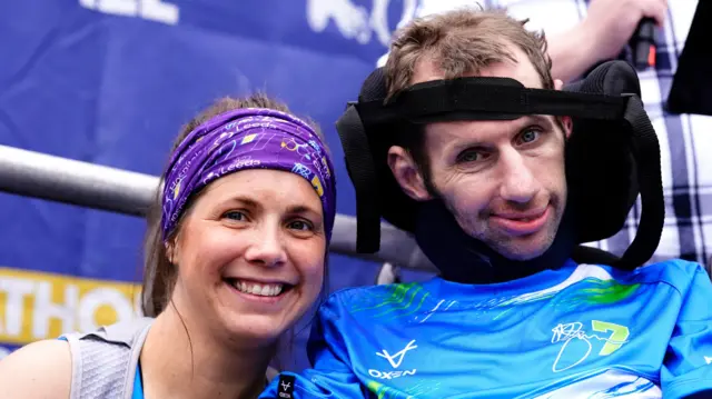 Lindsey and Rob Burrow. Lindsey is crouched down wearing a blue running top and blue and yellow headband. Rob is sat in a wheelchair, his head held in place by a supportive strap. They are both smiling.