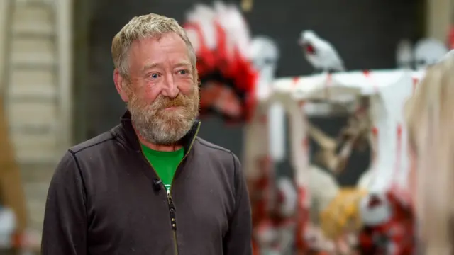 A man with a ginger beard and light hair smiles to the right of the camera, behind him are some Halloween decorationsdecorations