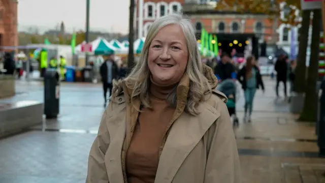 A woman in a brown coat smiles at the camera, she has long blonde hair and behind her are market stalls.
