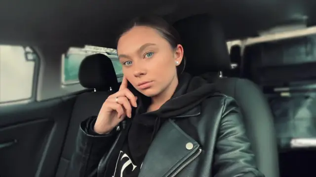 A selfie of Jaysley Beck in the front seat of a car with a black interior. She is wearing a black leather jacket and her hand is lightly touching her cheek.