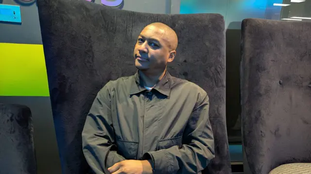 Rickie sat on a fabric chair with his arms crossed