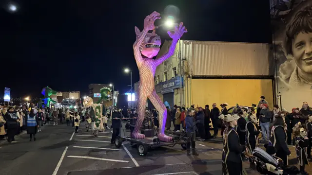A monster float is pulled along a street full of people