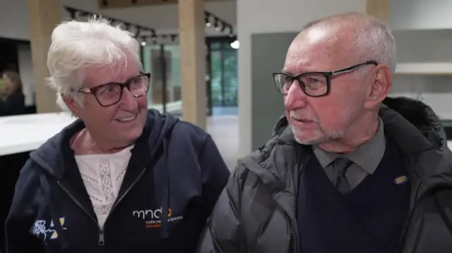 Irene and Geoff Burrow. Mrs Burrow is wearing a zip up MNDA hoodie over a white top. She has dark glasses and white hair. Mr Burrow is wearing a grey coat over a v-neck sweater, shirt and tie. He also has dark glasses, a grey beard and grey hair.
