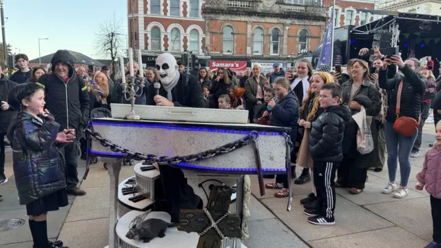 Man with his face painted white and black eyes is standing holding a microphone at a silver coffin made into a piano in a crowd of people