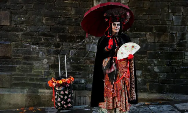 A woman in fancy dress is stood next to a suitcase - she has a decorative umbrella and a fan.
