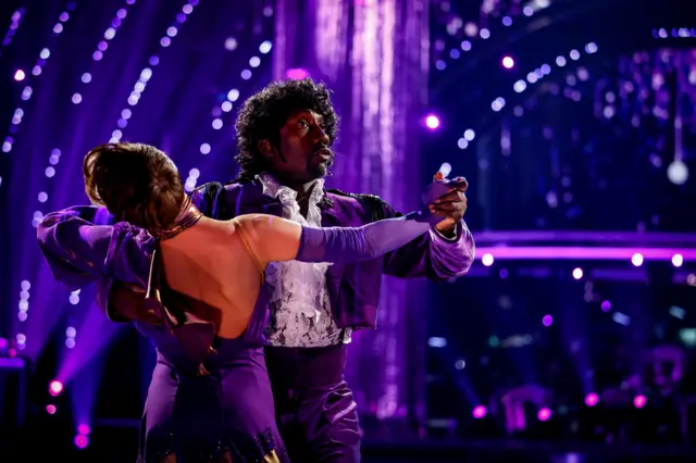 Jimmy is dressed in a purple sheen suit and wears a black wig, akin to the hairstyle that the late