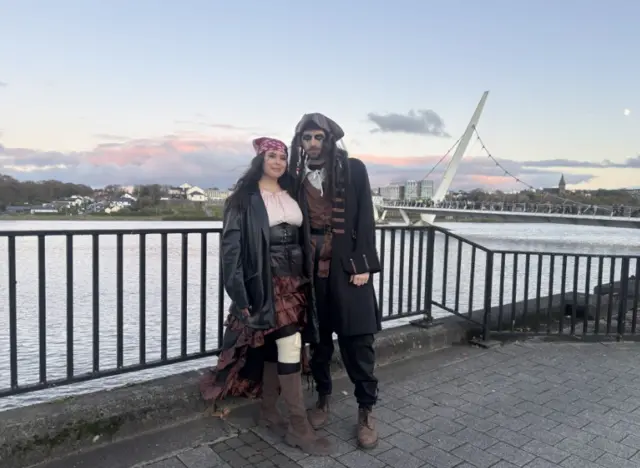 A man and a woman are dressed in pirate costumes, behind them is the Foyle Bridge