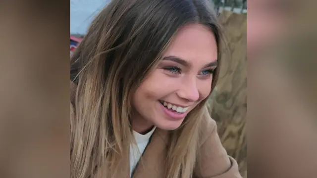 Jaysley Beck laughing and looking off to the left of the camera. She is wearing a brown blazer and has dark brown straight hair which is lighter towards the ends.