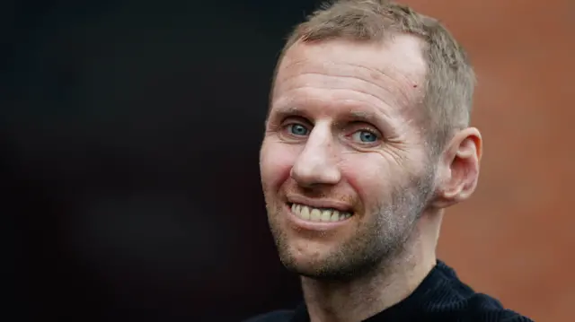 Rob Burrow smiles at the camera. He has short, light brown hair and blue eyes. He is wearing a dark coloured jumper