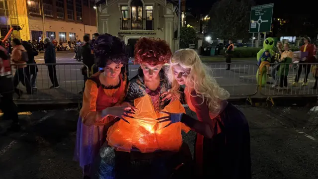 Three young girls are standing around a cauldron, with fabric that is made to look like a boiling cauldron
