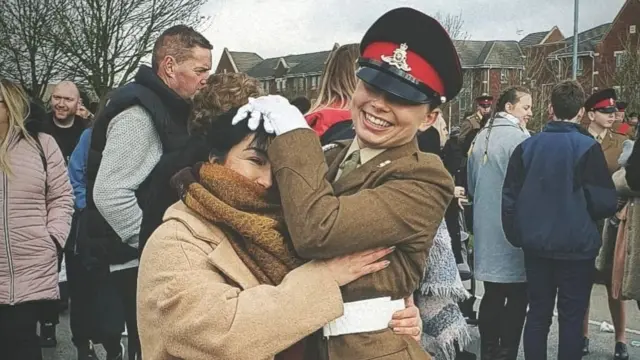 A young woman and her mother hug at a passing out ceremony. Jaysley is wearing a brown military uniform with a white belt and gloves and is playfully hugging her mum's head. There is a crowd of happy people milling about in the background.