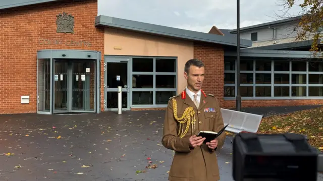 Major General Jon Swift standing outside the court, wearing a brown military uniform with gold ropes and red details.