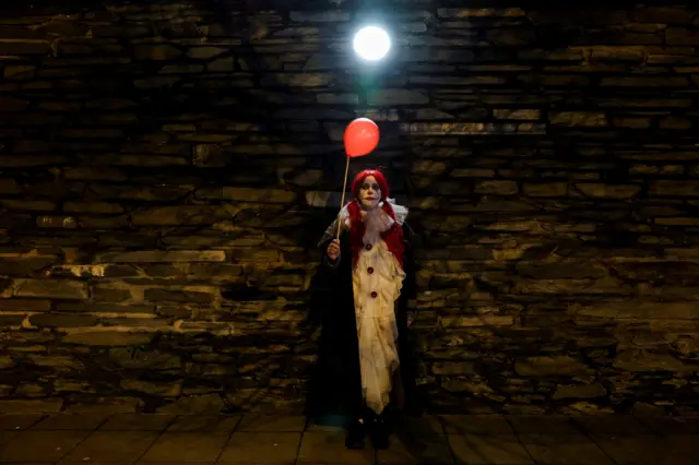 A girl dressed as It stands by a wall holding a red balloon