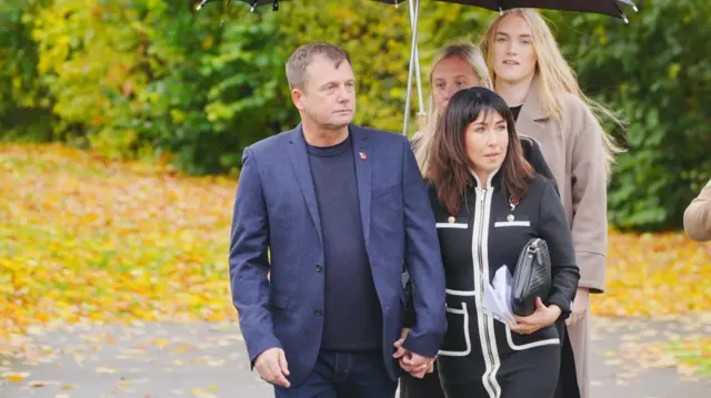 Gunner Beck's father Anthony Beck (L) and mother Leighann McCready (C) holding hands as they walk across a tarmac car park, with lots of autumnal leaves in the background.