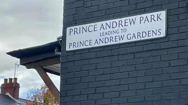 A white street sign reading 'Prince Andrew Park leading to Prince Andrew Gardens' on a grey brick wall