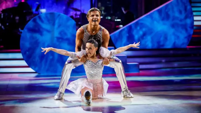 Amber Davies and Nikita wear silver outfits, with sparkly flourishes. He holds her as she does the splits between his legs, with her arms spread out