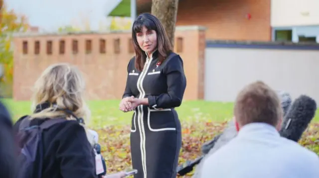 A woman in a smart black dress with white detailing stands and speaks to press. She is holding her hands in front of her and has a serious expression. The back of two members of the media can be seen.