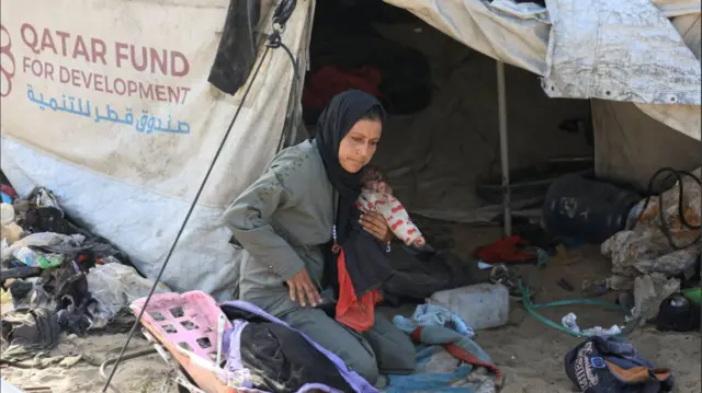 A woman holding a small child sits on her haunches outside a burlap tent, the sign Qatar Fund for Development on the side