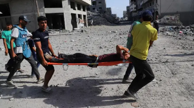 Three men run along a rubble street, carrying an injured man on a stretcher. There are flattened and bombed out buildings behind them.