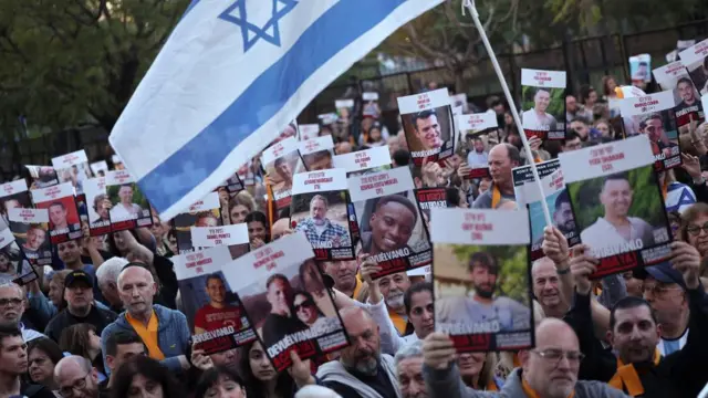 Dozens of people hold up signs of Israeli hostages, with one holding a large Israeli flag