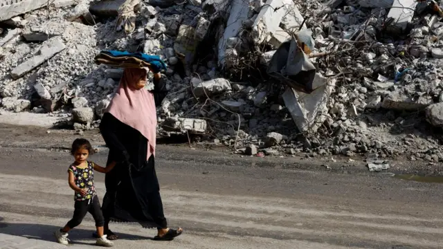 A Palestinian woman walks past a destroyed residential building, holding hands with a young girl