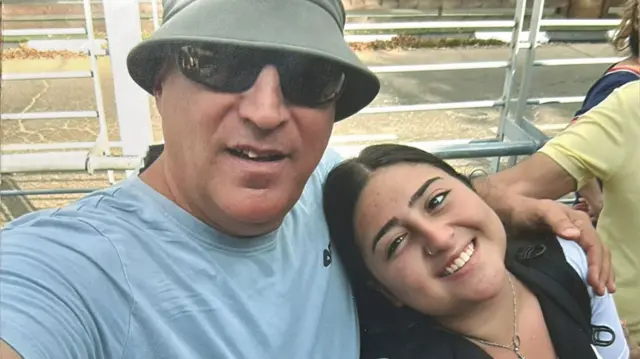Eyal Eshel wears a green bucket hat and blue t-shirt. he stands with his arm around his teen daughter roni, who has long dark hair. she is wearing a rucksack on her back and a silver necklace.