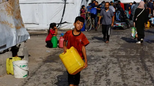 A displaced Palestinian boy carries a large container of water through a camp