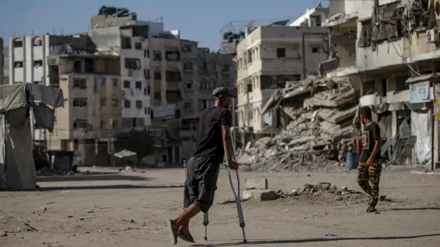 A man wearing a black t-shirt and a black cap walks on crutches, with destroyed building and rubble visible in the street ahead.