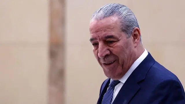 PA vice president Hussein al-Sheikh in a dark blue suit and patterned tie looks downwards to his left, a blurred beige wall behind him
