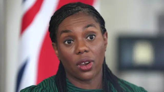 Kemi Badenoch head shot. She's mid-speech, wearing a green dress with black vertical stripes. There's a Union flag in background, blurred
