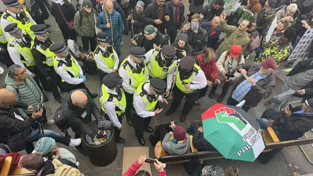 A group of Met Police officers in yellow hi-vi jackets approach a group of demonstrators holding up an umbrella in the Palestinian flag colours with the phrase Free Palestine on top during a protest