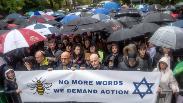 Dozens of people stand in the rain under umbrellas holding a sign which says "No more words, we demand action"