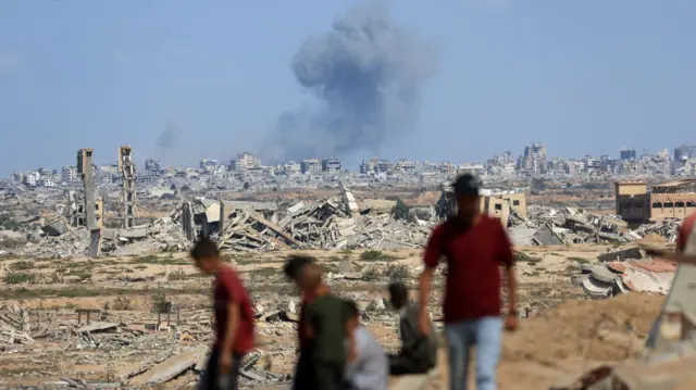 In the foreground: a group of six men and boys, blurred, stand as they observe billowing grey smoke rising into the sky after an air strike in gaza