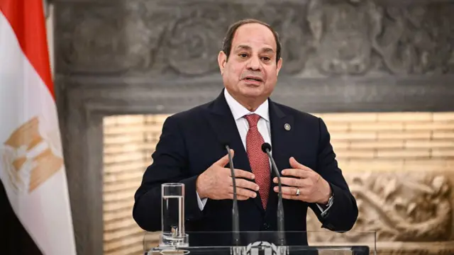 Egyptian President Abdel Fattah El-Sisi pictured in front of two microphones. He is wearing a black suit and red tie. There's an Egyptian flag in the background.