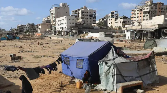 A woman crouches on the ground by a tent - city buildings can be seen behind her