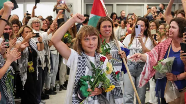 Greta raises her fist as she walks through a crowd of supporters at Athens airport. She is carrying flowers and a plaestinian flag can be seen partially obscured in the background.